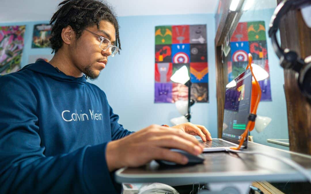 Young man wearing glasses and a blue hoodie working on a laptop at a desk, focused on his screen with colorful posters in the background.