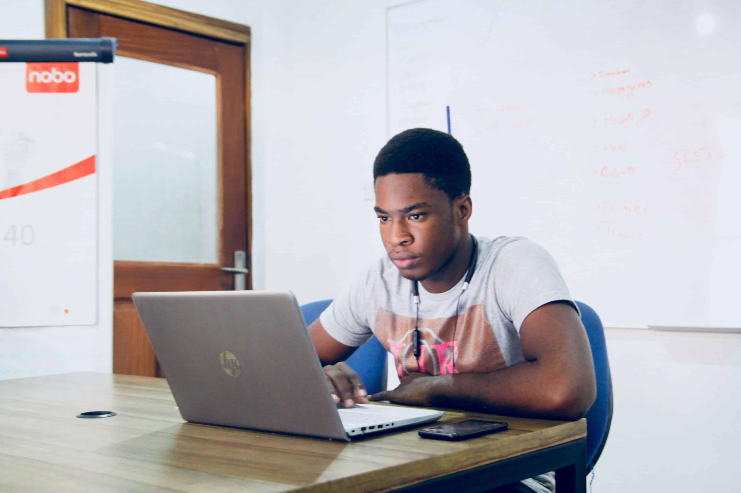Focused young man sitting at a desk working on a laptop, with notes written on a whiteboard in the background.