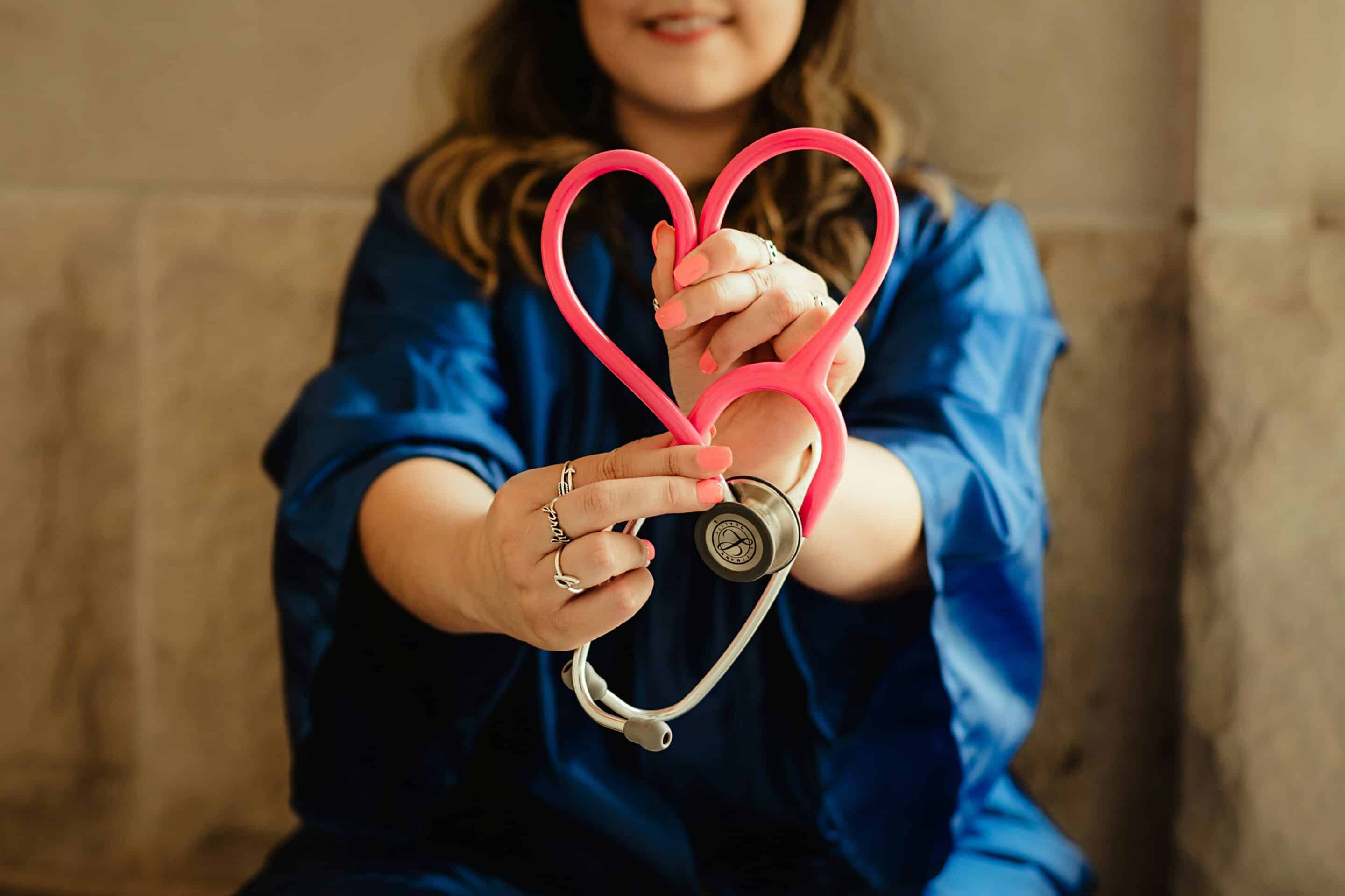 Woman in blue scrubs holding a pink stethoscope shaped like a heart, symbolizing care and compassion in healthcare.