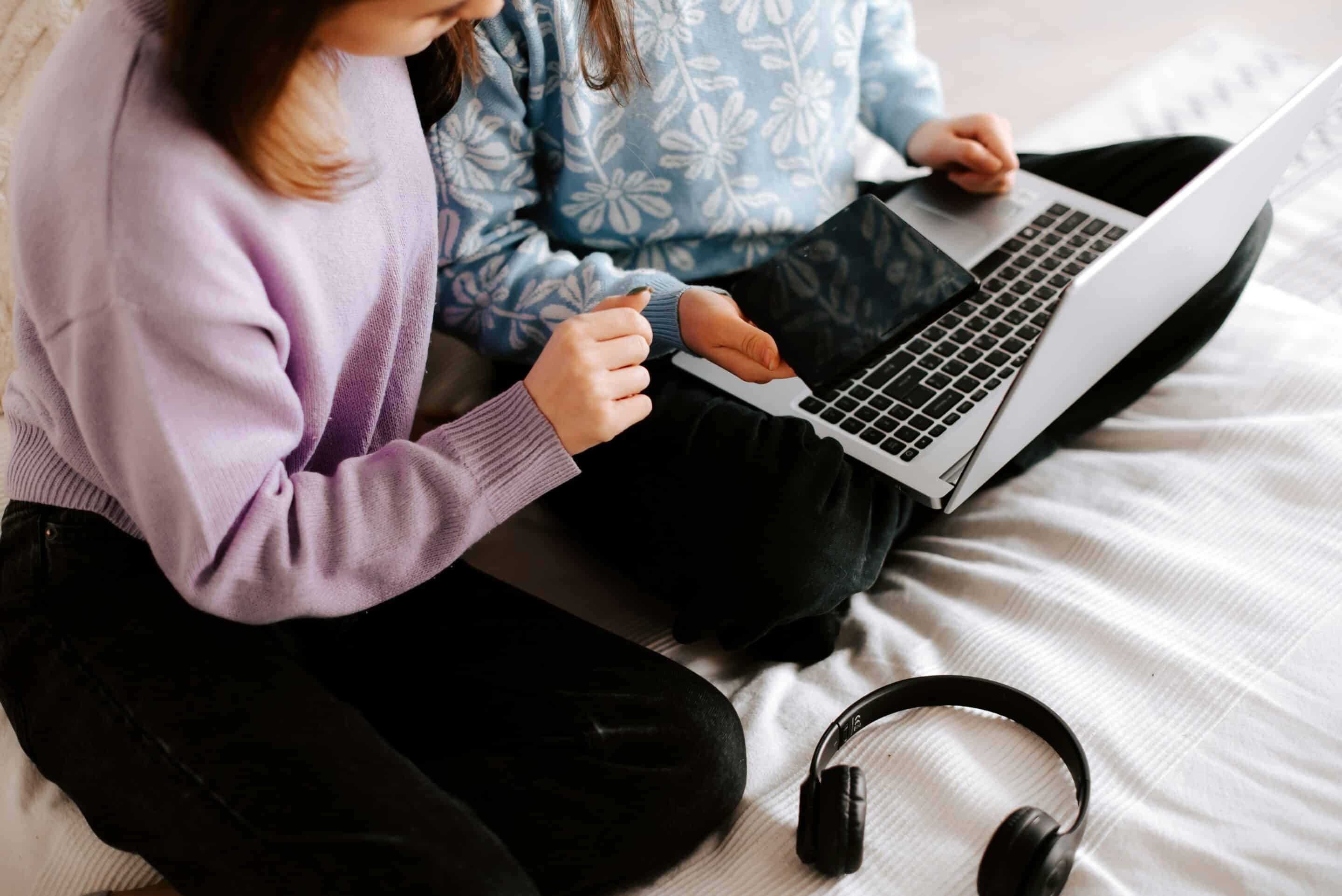 Two people sitting cross-legged on a bed, one holding a laptop and the other pointing at the screen, with headphones lying nearby.