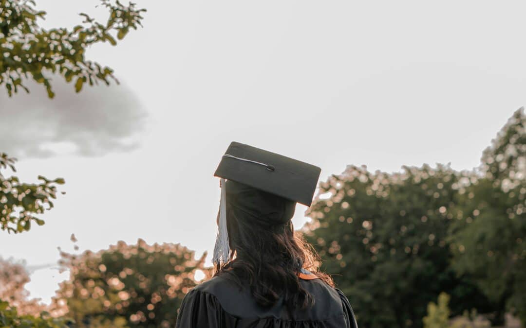 Graduate in cap and gown standing outdoors, facing the sunset with trees in the background, symbolizing looking forward to the future.