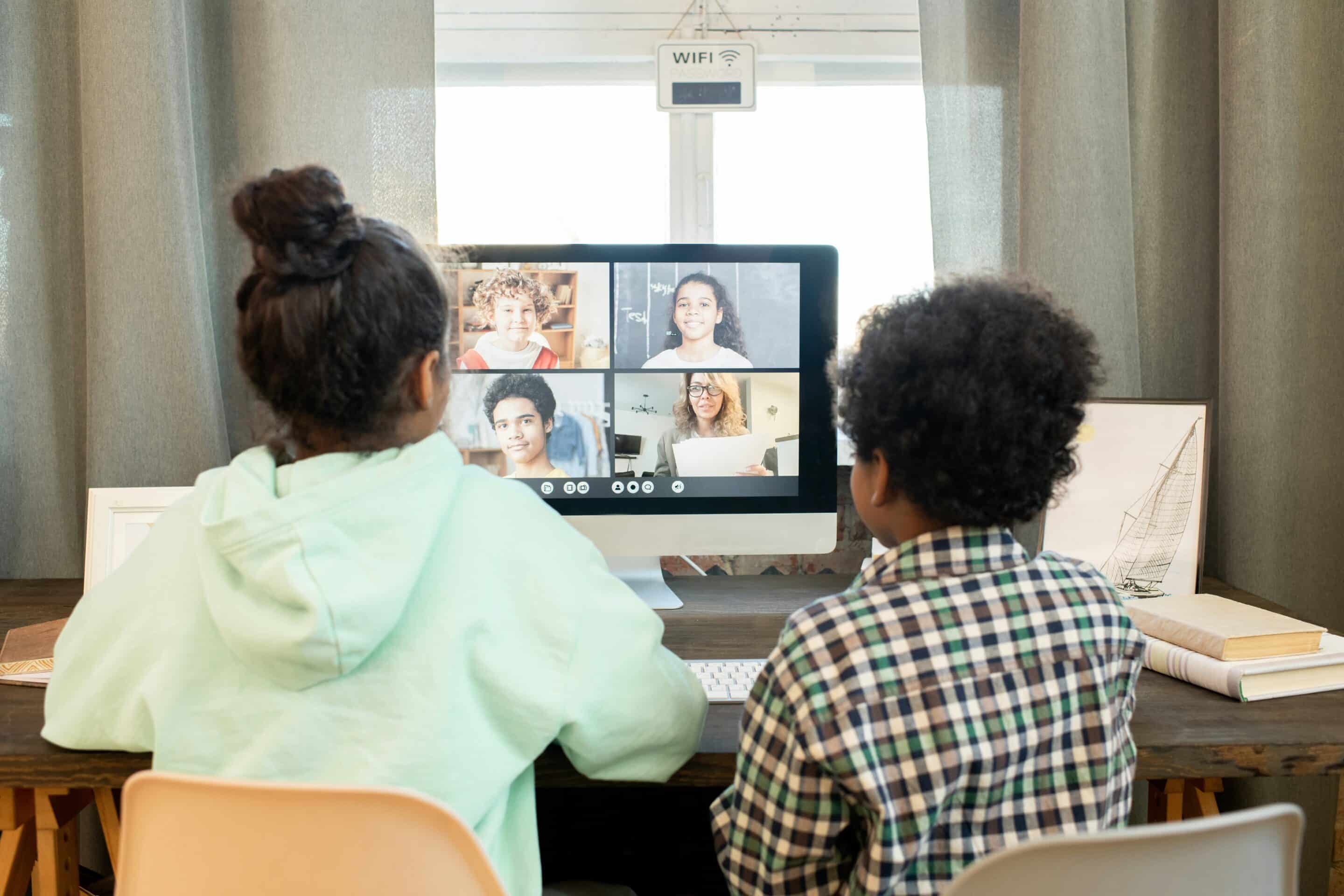 Two children sit at a desk and participate in an online class on a desktop computer, with four people visible on the video call screen, including other children and a teacher holding papers.