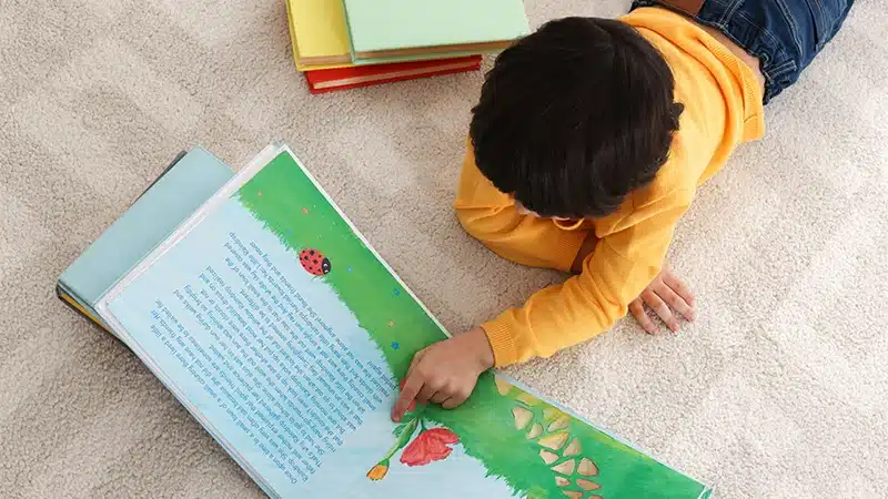 A young child with blonde hair sits at a wooden table, holding and reading a brightly colored “Hop on Pop” board book by Dr. Seuss. The child’s fingernails are painted pink, and soft natural light fills the room from a nearby window.